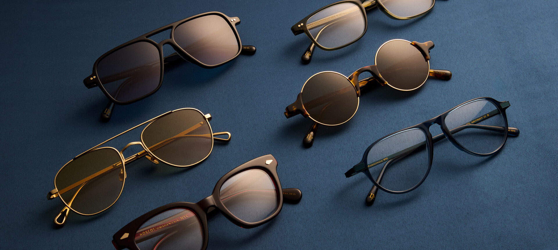 Seven pairs of Moscot glasses and sunglasses arranged on a dark blue surface, with MOSCOT/ORIGINALS/NYC and EST. 1915 visible on the temples