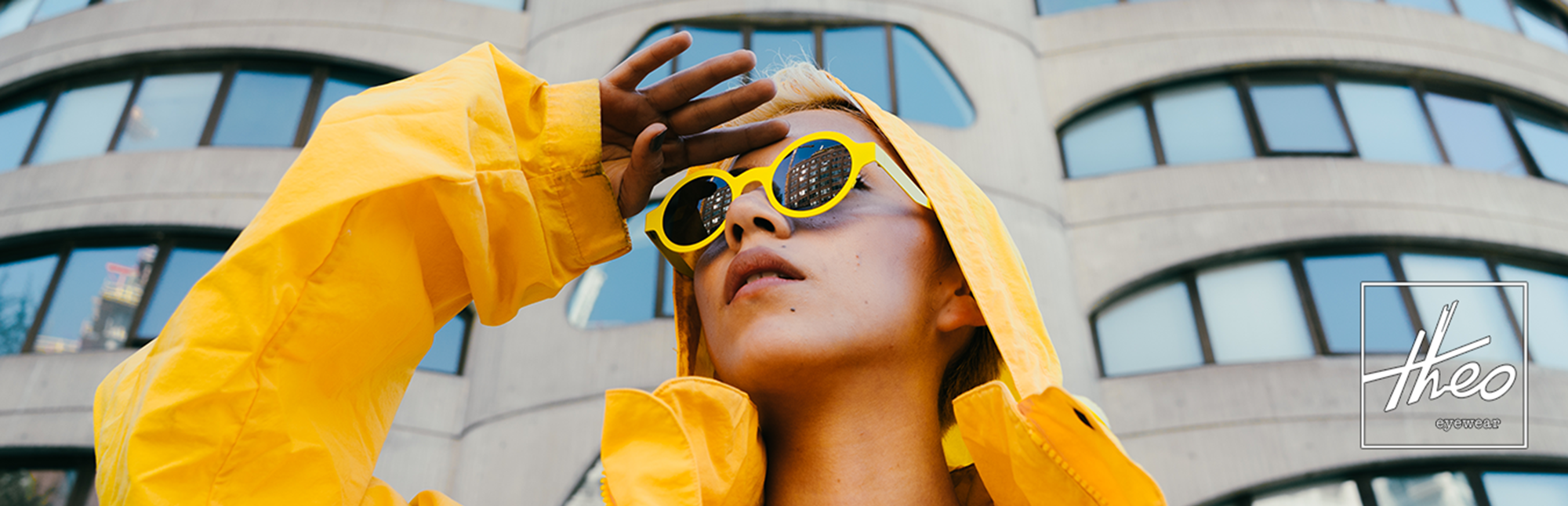 Person in a yellow raincoat wearing round yellow Theo sunglasses in front of a building